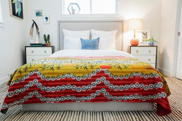 Best Bedsheets for Every Season and Comfort Needs 3 A modern bedroom with a white bed and a colorful patterned blanket in yellow and red tones, styled with bedside tables and lamps.