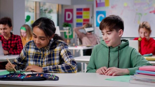 Two students sitting in a classroom, one drawing with colored pencils while the other looks on.
