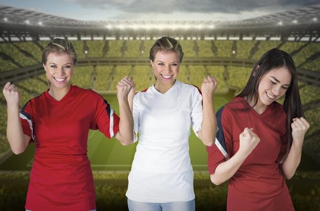 Three women in red and white soccer jerseys celebrating with clenched fists in a stadium background.
